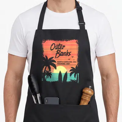 Tablier de cuisine noir avec illustration Outer Banks, palmiers, planches de surf et soleil couchant aux couleurs chaudes.