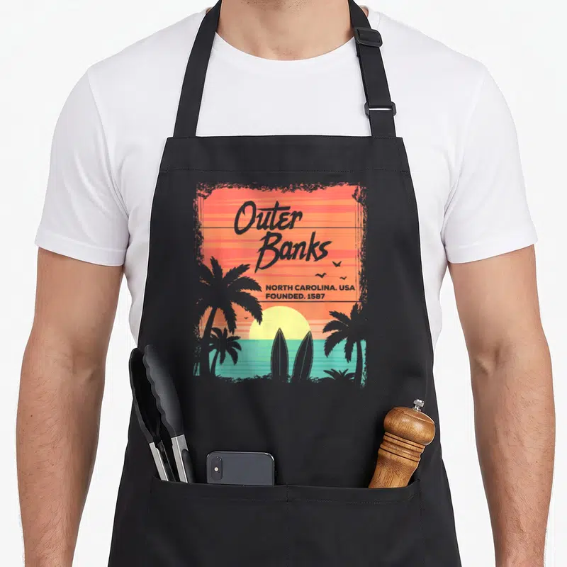 Tablier de cuisine noir avec illustration Outer Banks, palmiers, planches de surf et soleil couchant aux couleurs chaudes.