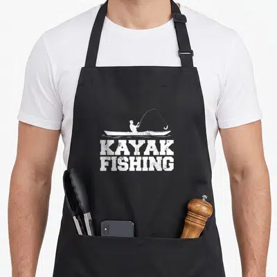 Tablier de cuisine noir avec silhouette blanche de pêcheur en kayak et texte KAYAK FISHING texturé.