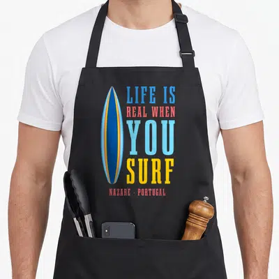 Tablier de cuisine noir avec planche de surf bleue et texte coloré Life is real when you surf Nazaré Portugal