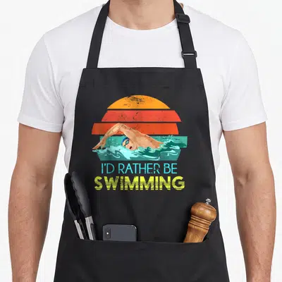 Tablier de cuisine noir avec illustration rétro d'un nageur effectuant un crawl sous un soleil orange et le texte I'd rather be swimming.