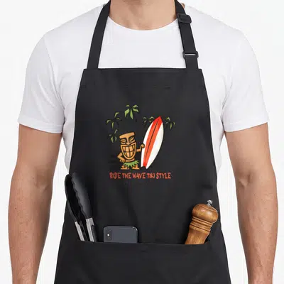 Tablier de cuisine noir avec un tiki souriant tenant une planche de surf orange et blanche sous des palmiers verts.