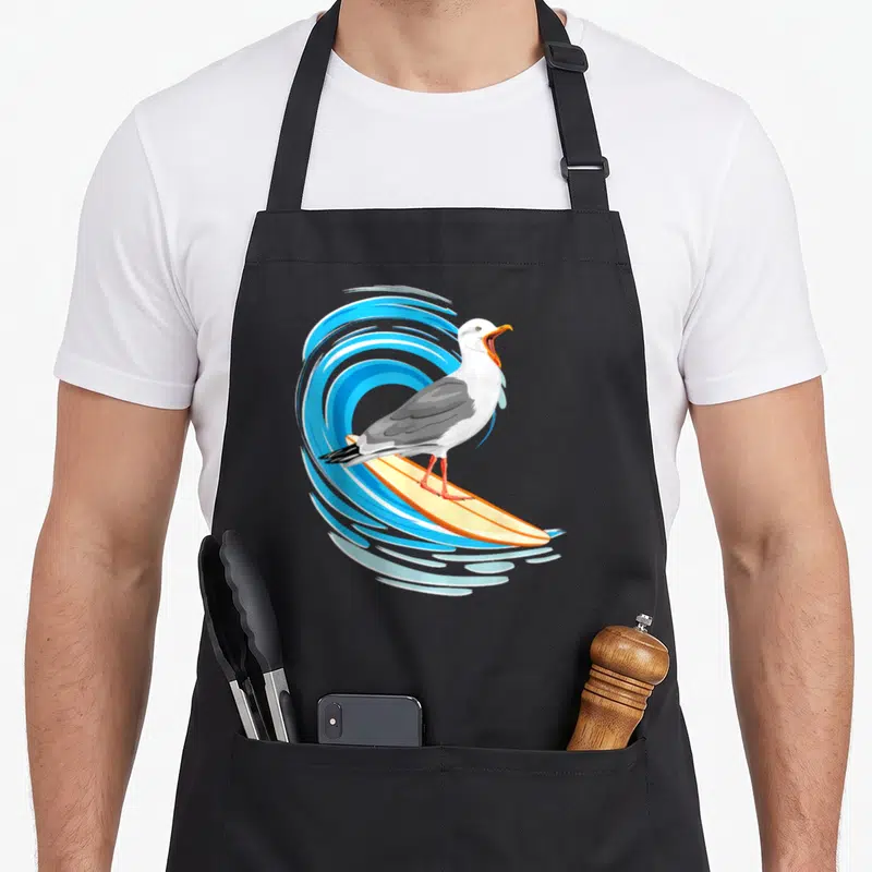 Tablier de cuisine noir avec mouette surfant sur planche jaune devant une vague bleue stylisée.