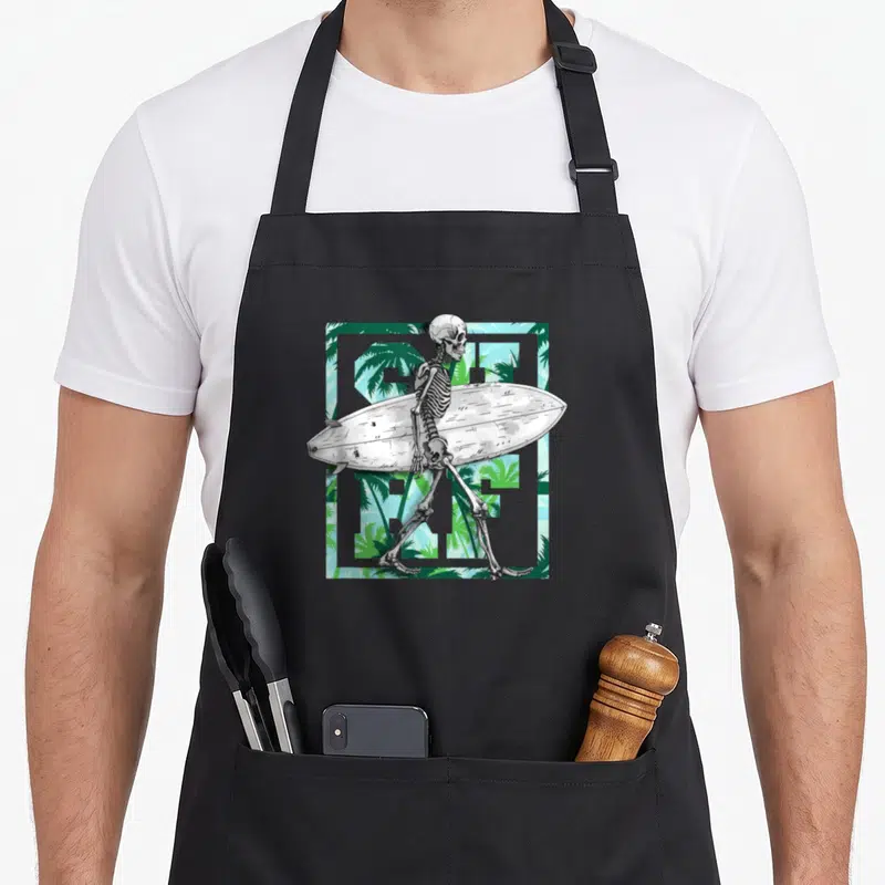 Tablier de cuisine noir avec un squelette tenant une planche de surf devant des palmiers verts.