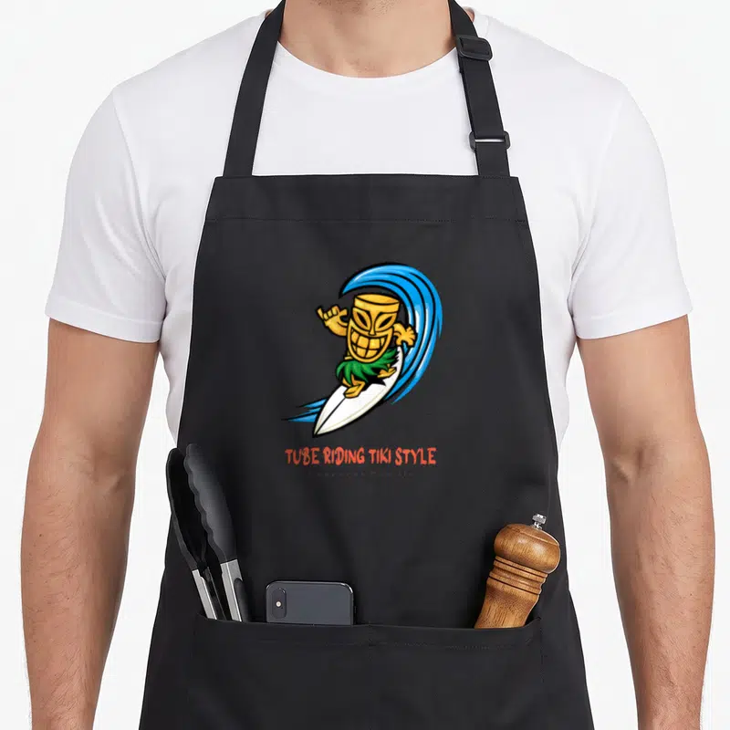 Tablier de cuisine noir avec un tiki souriant sur une planche de surf blanche et une vague bleue.