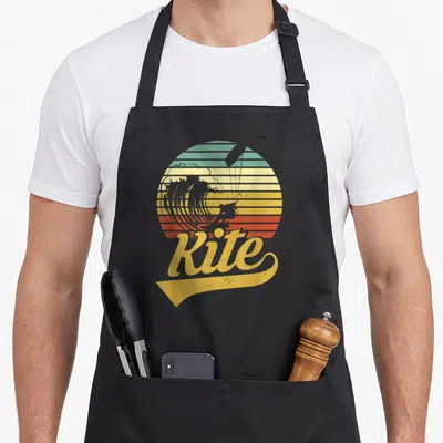 Tablier de cuisine noir avec illustration vintage d'un kitesurfeur sur vague et le mot Kite en jaune sous un soleil rayé.