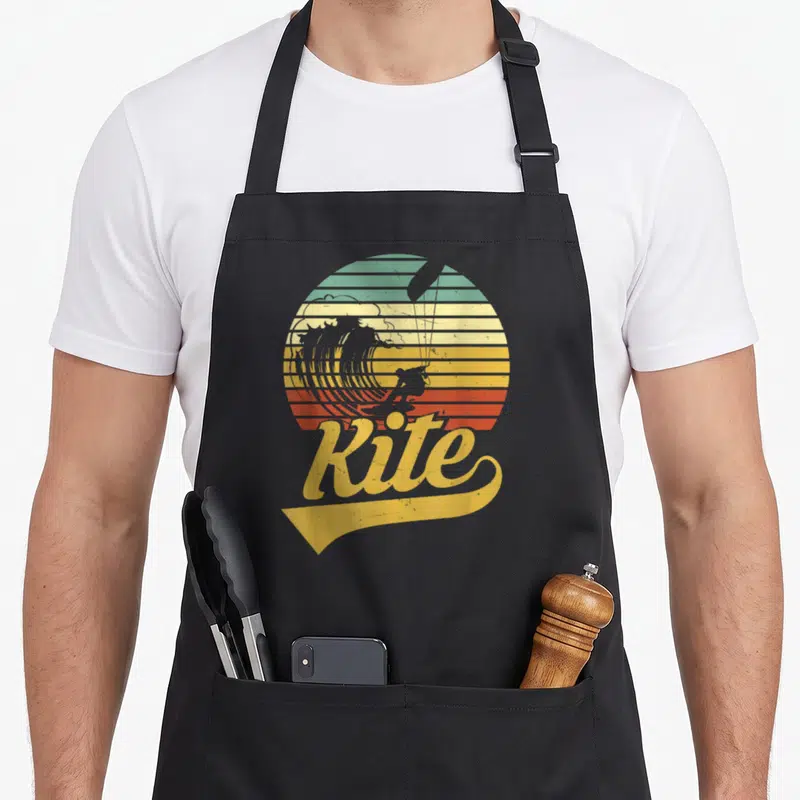 Tablier de cuisine noir avec illustration vintage d'un kitesurfeur sur vague et le mot Kite en jaune sous un soleil rayé.