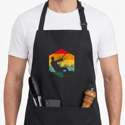 Tablier de cuisine noir avec un hexagone coloré représentant un kitesurfeur en silhouette grise sur fond rayé vintage.
