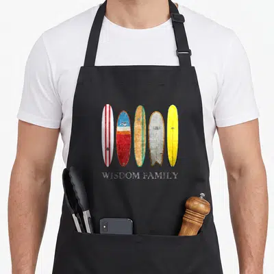 Tablier de cuisine noir avec cinq planches de surf vintage alignées et l'inscription Wisdom Family en bas.