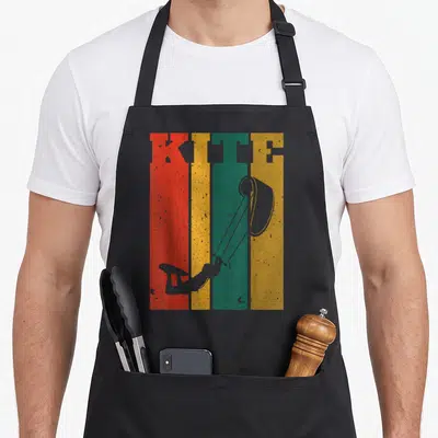 Tablier de cuisine noir avec illustration vintage de kitesurf sur bandes colorées et texte KITE en lettres usées.
