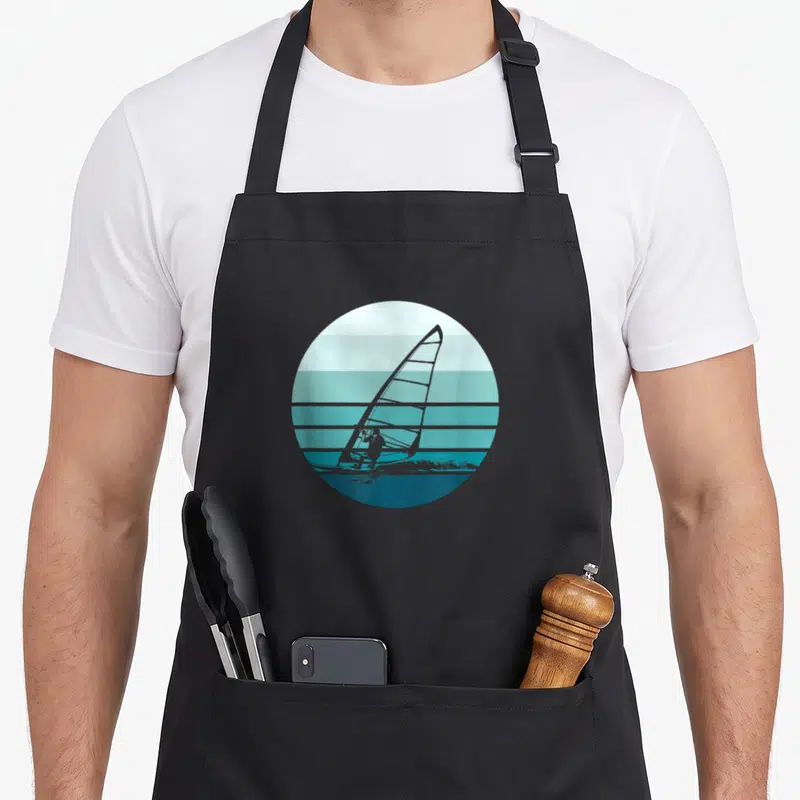 Tablier de cuisine noir avec illustration circulaire d'un windsurfeur sur fond de mer aux bandes turquoise et bleues.