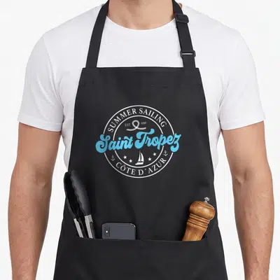 Tablier de cuisine noir avec logo circulaire Saint Tropez Summer Sailing en bleu et blanc incluant un voilier et des étoiles.
