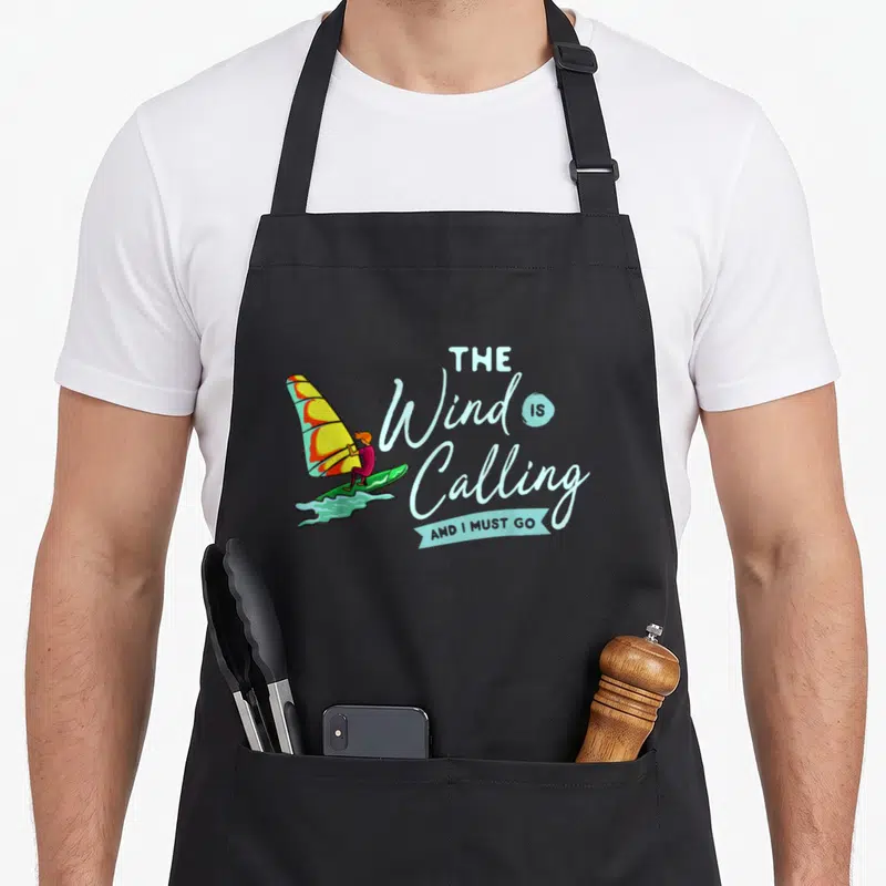 Tablier de cuisine noir avec illustration d'un windsurfer et texte The Wind Is Calling And I Must Go en bleu clair.
