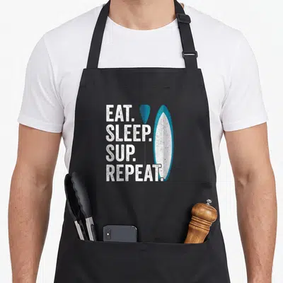 Tablier de cuisine noir avec l'inscription Eat Sleep SUP Repeat et une illustration de planche de paddle bleue et blanche.