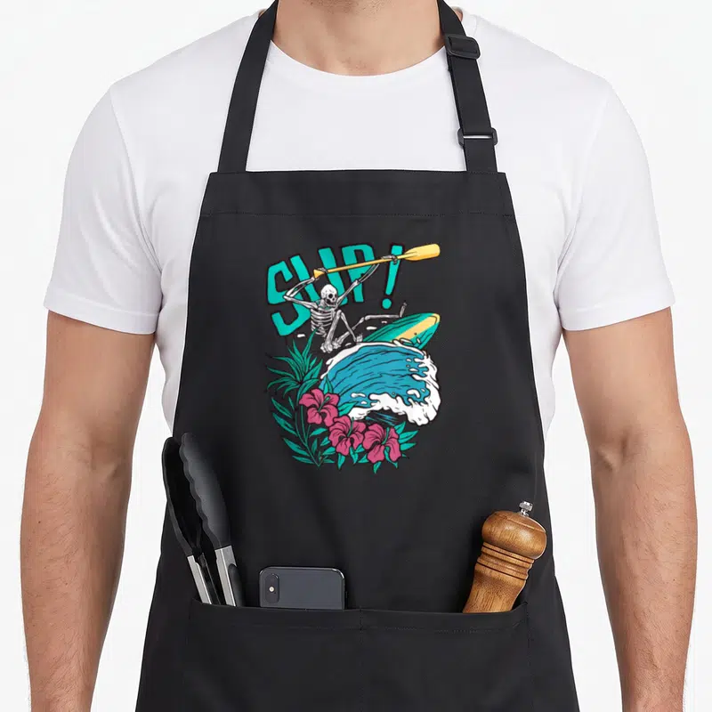 Tablier de cuisine noir avec un squelette en stand up paddle sur une vague et des fleurs tropicales roses.