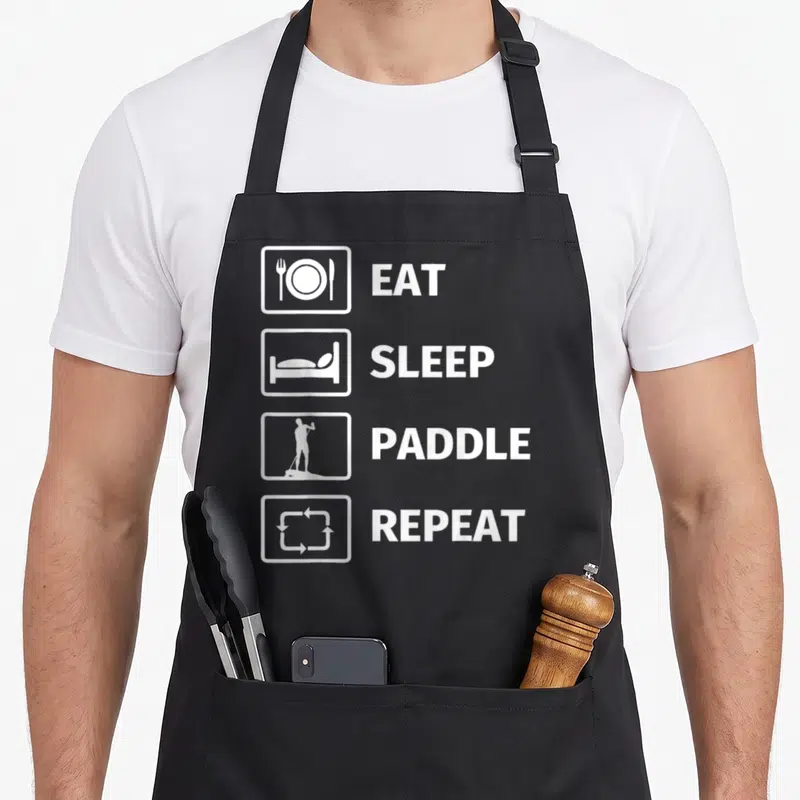 Tablier de cuisine noir avec quatre icônes blanches verticales et les mots Mangez Sleep Paddle Repeat imprimés en majuscules.