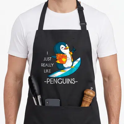 Tablier de cuisine noir avec un pingouin surfeur en chemise orange et le texte I Just Really Like Penguins.