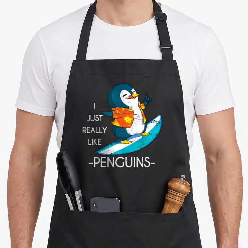 Tablier de cuisine noir avec un pingouin surfeur en chemise orange et le texte I Just Really Like Penguins.