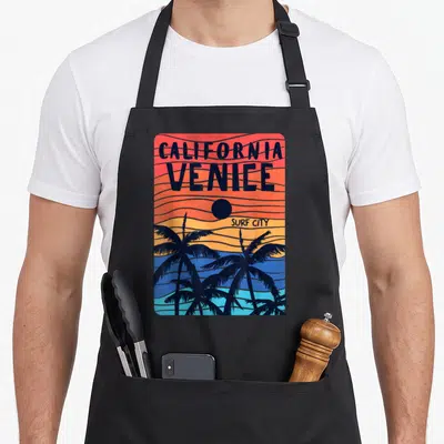 Tablier de cuisine noir avec illustration rétro de palmiers et texte California Venice Surf City sur fond dégradé orange et bleu.
