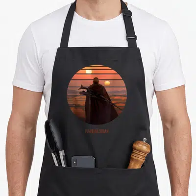 Tablier de cuisine noir avec illustration circulaire du Mandalorien dos et fusil sous un soleil orange stylisé avec texte The Mandalorian en bas.