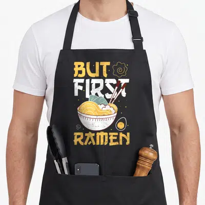 Tablier de cuisine noir avec illustration d'un bol de ramen, œuf, baguettes et texte But First Ramen en jaune et blanc.
