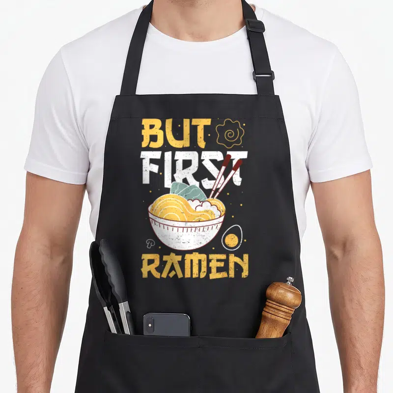 Tablier de cuisine noir avec illustration d'un bol de ramen, œuf, baguettes et texte But First Ramen en jaune et blanc.