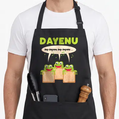 Tablier de cuisine noir avec trois grenouilles vertes lisant des parchemins et chantant Dayenu sous un nuage de notes musicales.