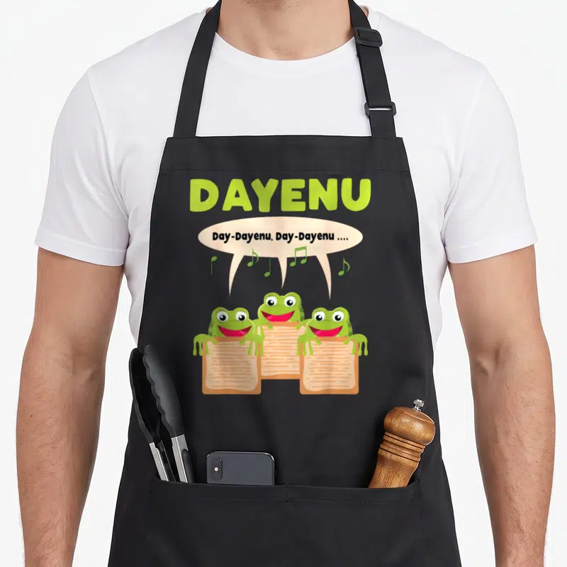 Tablier de cuisine noir avec trois grenouilles vertes lisant des parchemins et chantant Dayenu sous un nuage de notes musicales.