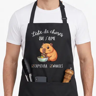 Tablier de cuisine noir avec illustration d'un capybara mangeant des nouilles dans un bol blanc et vert.