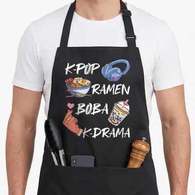 Tablier de cuisine noir avec illustration de bol de ramen, casque audio, gobelet à boba et geste du cœur en blanc sur fond uni.