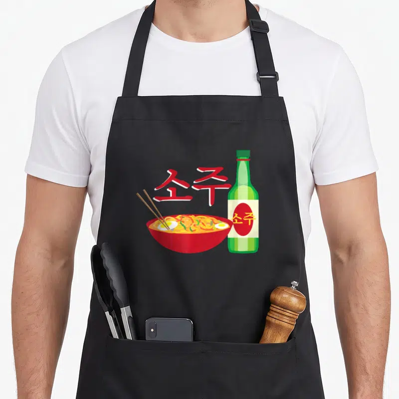 Tablier de cuisine noir avec illustration d'un bol de ramen rouge, d'une bouteille verte de soju et de caractères coréens rouges stylisés.