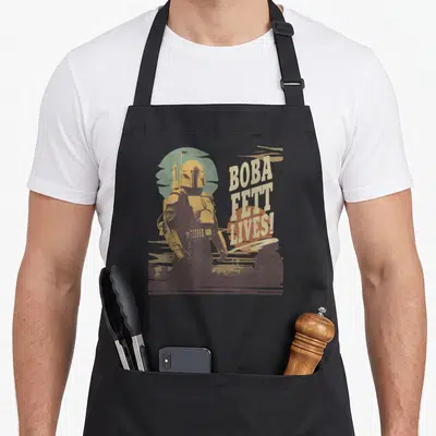 Tablier de cuisine noir avec une illustration rétro de Boba Fett et le texte « BOBA FETT LIVES! » en lettres capitales beige et orange.