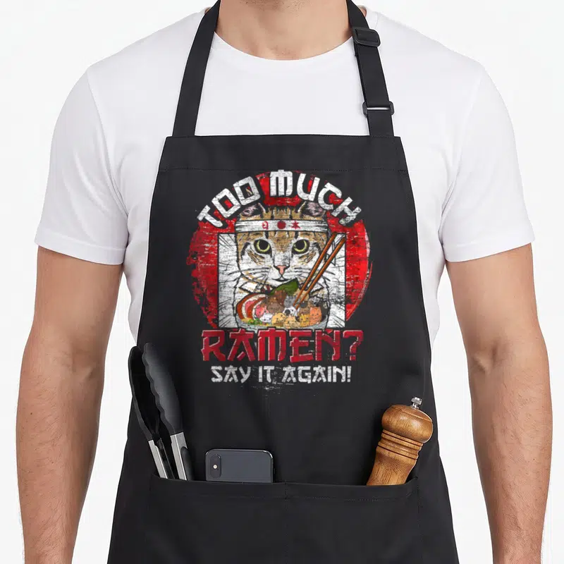 Tablier de cuisine noir avec un chat samouraï, des nouilles ramen et les mots Too Much Ramen Say It Again imprimés en rouge et blanc.