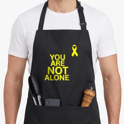 Tablier de cuisine noir avec texte jaune You Are Not Alone et ruban de solidarité en haut à droite.