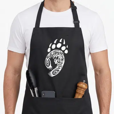 Tablier de cuisine noir avec une illustration blanche représentant une patte d'ours et un visage tribal grizzly au centre.