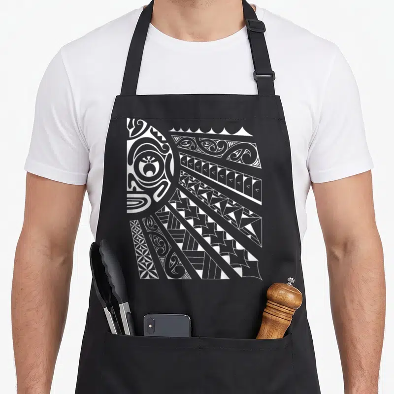 Tablier de cuisine noir avec un grand motif tiki soleil polynésien en blanc aux lignes tribales complexes.