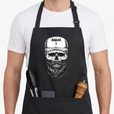 Tablier de cuisine noir avec illustration d'un crâne barbu au casque et texte Barbe & Tatouages CULTE en typographie vintage.