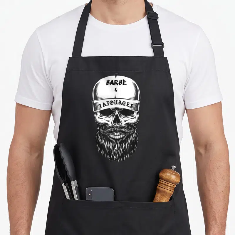 Tablier de cuisine noir avec illustration d'un crâne barbu au casque et texte Barbe & Tatouages CULTE en typographie vintage.