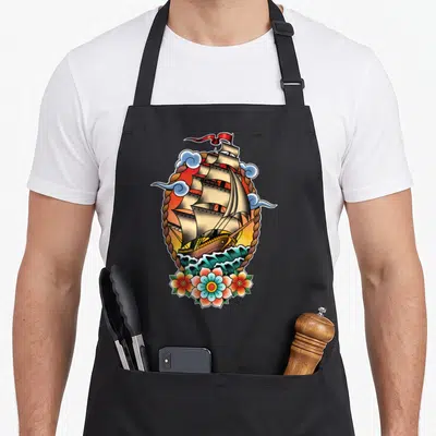 Tablier de cuisine noir avec illustration navire old school, cordes tressées, nuages bleus et fleurs colorées en bas du motif.
