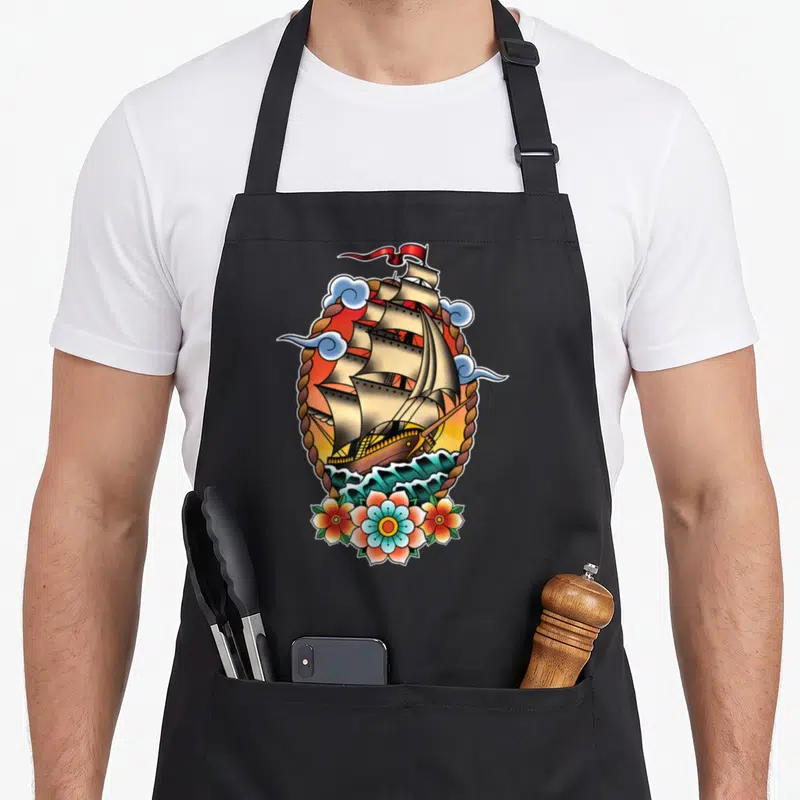 Tablier de cuisine noir avec illustration navire old school, cordes tressées, nuages bleus et fleurs colorées en bas du motif.