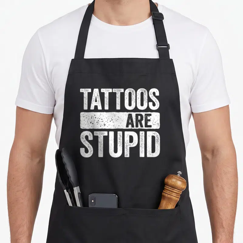 Tablier de cuisine noir avec l'inscription blanche Tattoos Are Stupid en lettres capitales texturées.
