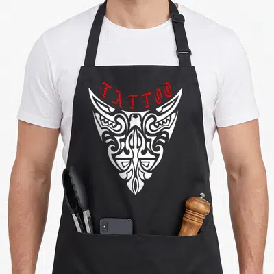 Tablier de cuisine noir avec un grand motif tribal blanc et le mot Tattoo écrit en rouge au sommet.