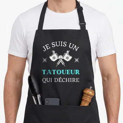 Tablier de cuisine noir avec deux machines à tatouer croisées et le texte Je suis un tatoueur qui déchire en blanc et bleu cyan.
