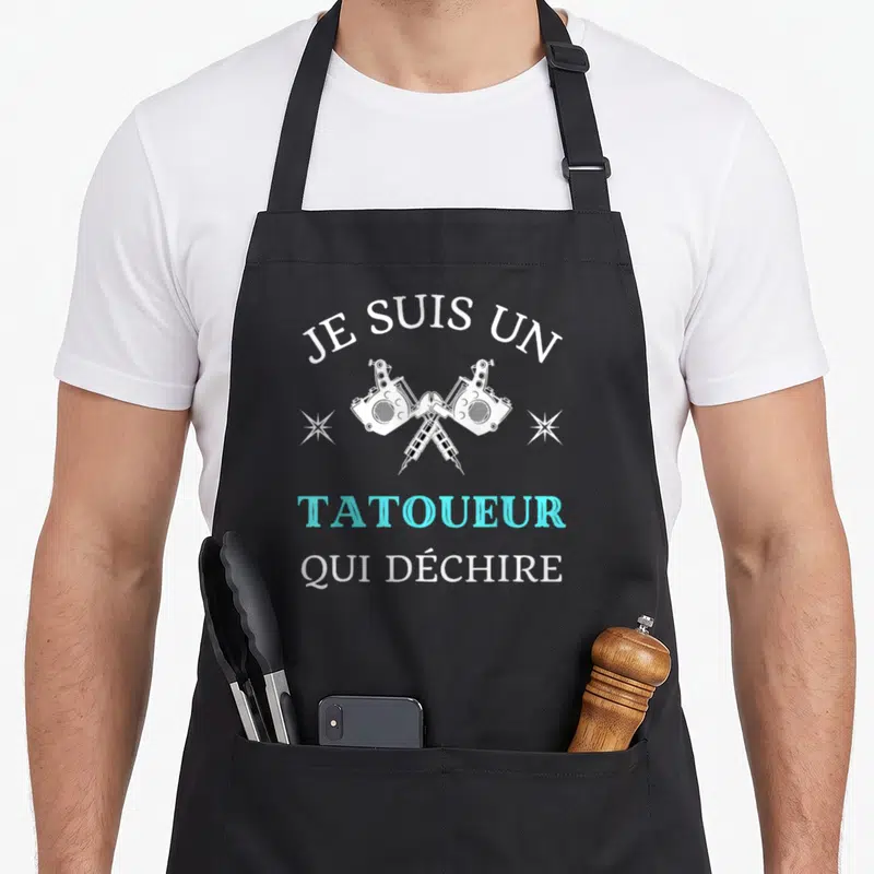 Tablier de cuisine noir avec deux machines à tatouer croisées et le texte Je suis un tatoueur qui déchire en blanc et bleu cyan.