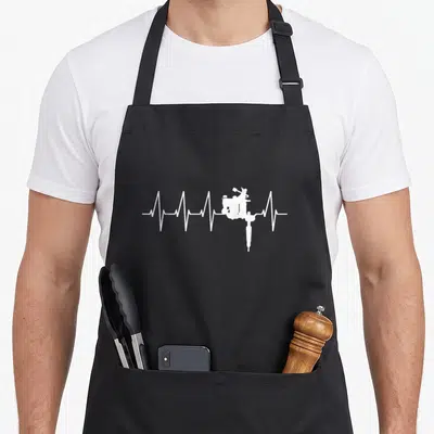 Tablier de cuisine noir avec motif blanc en forme de battement de cœur intégrant une machine à tatouer.