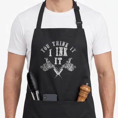Tablier de cuisine noir avec inscription blanche You Think It I Ink It et trois machines à tatouer dessinées en blanc.