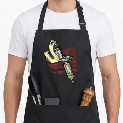 Tablier de cuisine noir avec illustration colorée d'une machine à tatouer jaune et argentée sur fond rouge texturé.