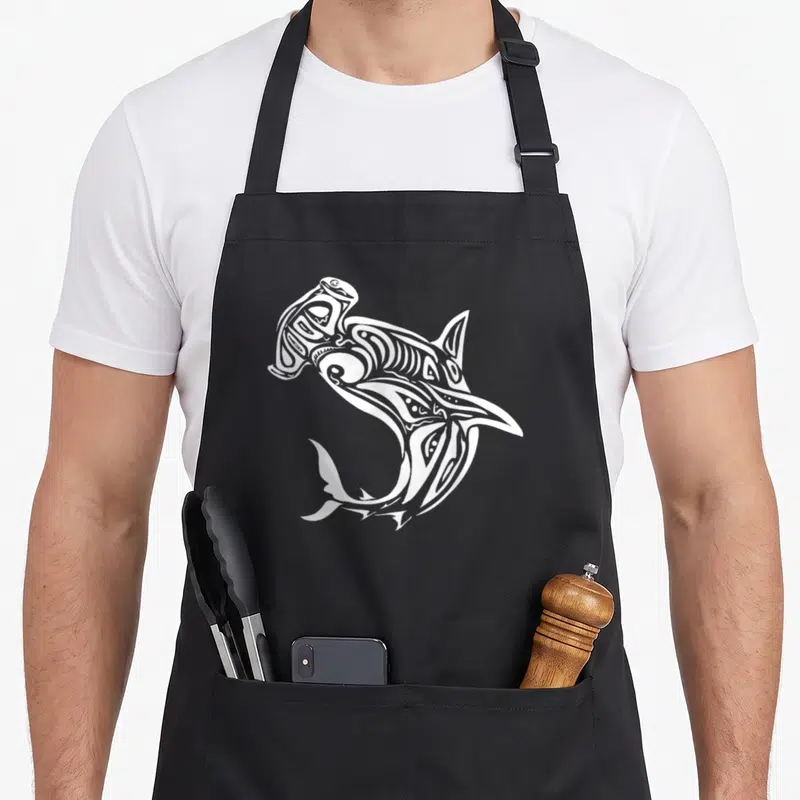 Tablier de cuisine noir avec un requin polynésien Maori tribal imprimé en blanc au centre.