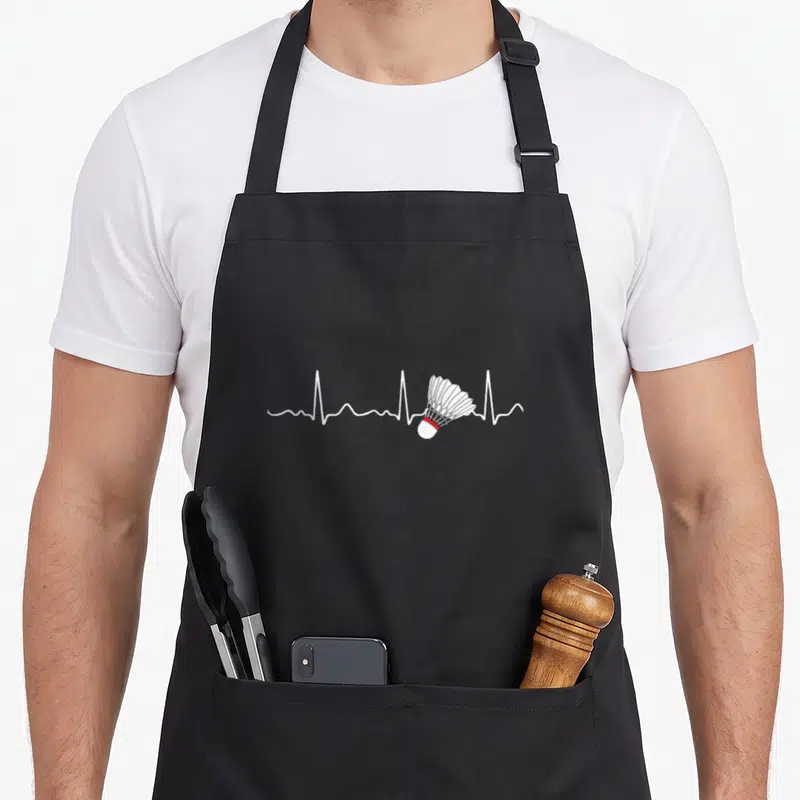 Tablier de cuisine noir avec ligne blanche de rythme cardiaque intégrant un volant de badminton au centre du dessin.
