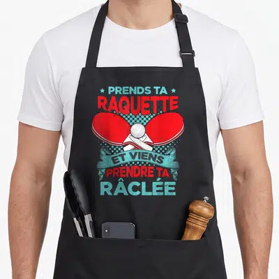 Tablier de cuisine noir avec deux raquettes de ping-pong rouges croisées et un ballon blanc au centre.